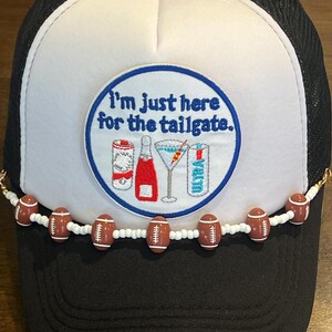 May include: Black and white trucker hat with a white front panel and a mesh back. The hat features a circular patch with the text "I'm just here for the tailgate." A beaded chain of footballs adorns the brim.