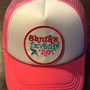 Op de afbeelding: Een roze truckerpet met een wit voorpaneel en een roze mesh achterkant. De pet heeft een cirkelvormige patch met de tekst "Santa's favorite ho" in rood, teal en wit, met een hulsttakje. De klep en de bovenste knop zijn ook roze.