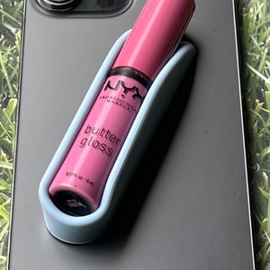 Puede incluir: Un tubo de brillo labial rosa con el texto "NYX PROFESSIONAL MAKEUP butter gloss 0.27 fl oz / 8 ml" en la etiqueta. El tubo está en un agarre de teléfono azul claro.