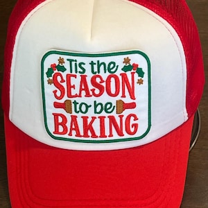May include: Red and white trucker hat with a mesh back. The front panel is white with a rectangular patch that reads "Tis the SEASON to be BAKING" in red and green. A festive accessory for baking enthusiasts.