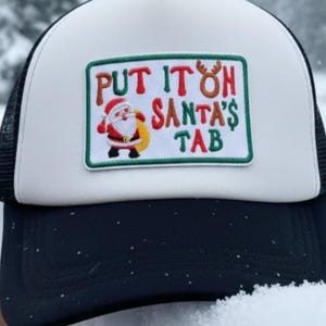 May include: A black and white trucker hat with a white front panel and a black mesh back. The hat features a patch with the text "PUT IT ON SANTA'S TAB" and a cartoon Santa Claus.