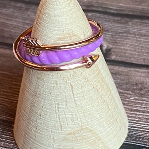 May include: A purple and gold adjustable ring with an arrow design. The ring is on a wooden display stand.