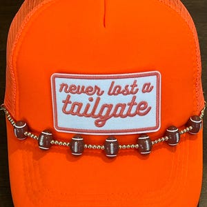 May include: Bright orange trucker hat with a mesh back and a curved brim. The hat features a patch with the text "never lost a tailgate" and a beaded football accent.