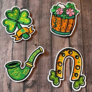 May include: Four embroidered patches with a St. Patrick's Day theme. The patches include a green shamrock, a wooden barrel with flowers, a green pipe, and a yellow horseshoe with green shamrocks.