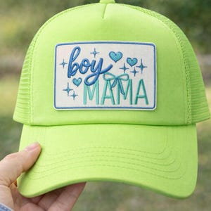 May include: A neon green trucker hat with a rectangular patch. The patch features the words "boy MAMA" in blue and teal embroidery, along with hearts and stars. The hat has a mesh back and a curved brim.