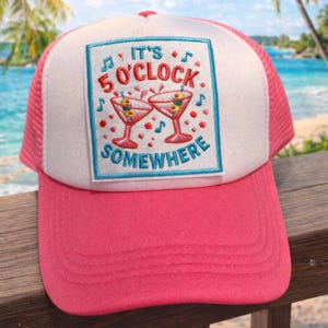May include: A pink and white trucker hat featuring the text "It's 5 O'Clock Somewhere" with cocktail glasses. The hat has a pink peak and mesh back, ideal for a day at the beach or a summer holiday.