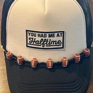 May include: A black and white trucker hat with the text "YOU HAD ME AT Halftime" on the front. The hat features a beaded chain of football-shaped beads along the brim. The hat has a mesh back.