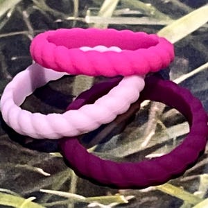 May include: Three silicone rings in shades of pink and purple. The rings have a braided rope design. The colors include a bright pink, a light pink, and a deep purple. The rings are stacked on top of each other.