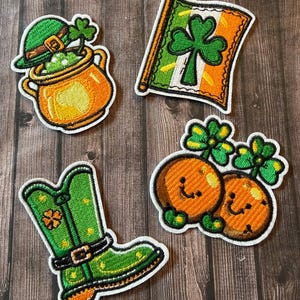 May include: Four embroidered patches with St. Patrick's Day themes. Includes a pot of gold with a leprechaun hat, an Irish flag with a shamrock, a green boot with a buckle, and two orange smiling characters with shamrocks.