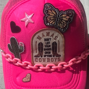 May include: Pink trucker hat with a variety of patches, including a butterfly, a star, a cactus, and a heart. The hat also has a pink chain around the brim and a patch that says "Mama's Don't Let Your Babies Grow Up To Be Cowboys".