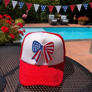 May include: A red and white trucker hat featuring a central embroidered patch of a bow. The bow design incorporates a flag pattern with white stars on a blue field on the left side and red and white stripes on the right side of the bow.