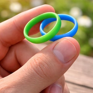 Könnte beinhalten: Zwei Silikonringe, einer in leuchtendem Grün und einer in leuchtendem Blau, werden zwischen Fingern gehalten. Die Ringe sind glatt und haben eine matte Oberfläche. Der Hintergrund ist verschwommen, mit Andeutungen von grünem Laub und Sonnenlicht.