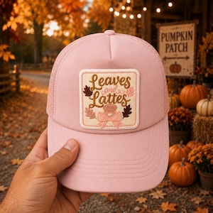 May include: A light pink trucker hat featuring a square embroidered patch on the front. The patch displays the text Leaves and Lattes in brown script, surrounded by small embroidered autumn leaves and a coffee cup graphic with a pink bow.