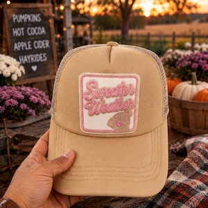 May include: A tan trucker hat featuring a square patch on the front. The patch has a pink embroidered border and the text "Sweater Weather" in pink script, accompanied by a small embroidered graphic of a knit beanie with a pink heart detail.