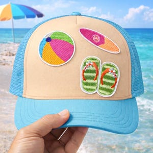 May include: A light blue and tan trucker hat with a beach theme. The hat features embroidered patches of a beach ball, surfboard, and flip-flops. The hat has a blue brim and mesh backing, perfect for a day at the beach.