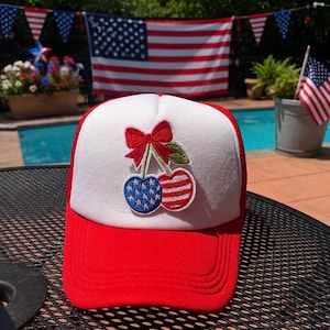 May include: A white and red trucker hat featuring an embroidered patch of two cherries tied with a red bow. One cherry is blue with white stars, and the other cherry has red and white horizontal stripes, mimicking the American flag design.