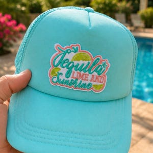 Puede incluir: Gorra de camionero turquesa con parte trasera de malla y visera curva. La gorra presenta un parche bordado con las palabras "Tequila Lime and Sunshine" en una fuente lúdica, con gráficos de lima y corazón.