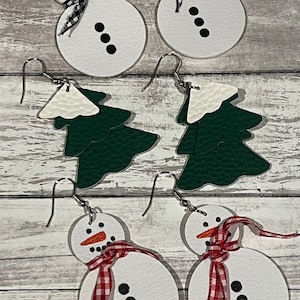 May include: Four pairs of Christmas-themed earrings. Two pairs feature white snowman ornaments with black eyes, red noses, and black and white or red and white ribbon accents. The other two pairs feature green Christmas tree ornaments with white snow caps.