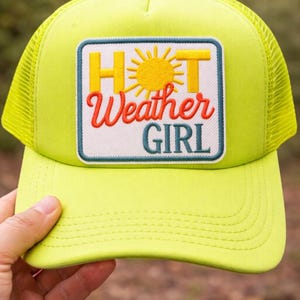 May include: A neon yellow trucker hat with a mesh back. The front features a patch with the words "HOT Weather GIRL" and a sun graphic. The hat is a bright, eye-catching accessory.