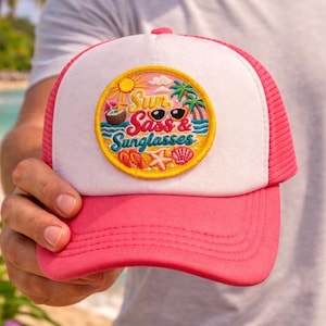 以下が含まれることがあります： 「Sun, Sass & Sunglasses」の文字が入った円形パッチが付いた、ピンクと白のトラッカーハット。パッチにはサングラス、ヤシの木、ココナッツドリンクのイラストが含まれています。帽子は人が持っています。
