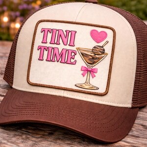 May include: Brown and beige trucker cap with a patch reading "TINI TIME" in pink. The patch shows a pink heart and a cocktail with a chocolate-covered ball. The cap has a brown peak and mesh back.