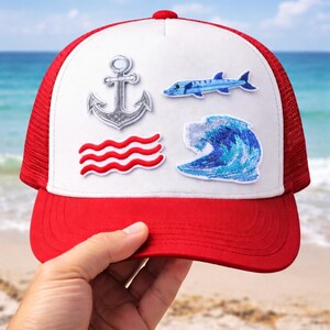 Peut inclure: Casquette de camionneur rouge et blanche avec des écussons brodés d'une ancre, de vagues, d'un poisson et d'une vague. La casquette a une visière rouge et un dos en filet, parfaite pour une journée à la plage.
