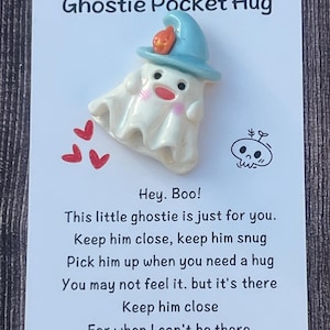 May include: A white ceramic ghost figurine with a blue hat and orange flame detail, labeled "Ghostie Pocket Hug." The card includes the text "Hey. Boo!" and a poem about keeping the ghost close for comfort.