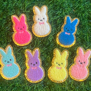 May include: Seven colourful felt bunny shaped patches with gold glitter trim. The bunnies are pink, blue, white, yellow, purple, and light blue.