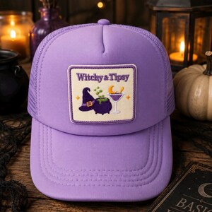 Puede incluir: Una gorra tipo trucker de color morado claro con un parche rectangular en la parte frontal. El parche muestra el texto Witchy & Tipsy en morado, junto a un gráfico bordado de un sombrero de bruja morado, un caldero negro y una copa de martini.