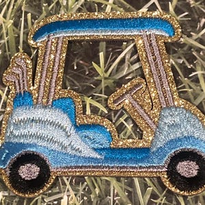 May include: A blue and gold glitter golf cart patch. The cart has a gold glitter roof and a gold glitter steering wheel. The cart is on a green grass background.