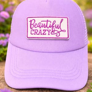 May include: A lavender trucker hat with a curved brim and mesh back. The front features a rectangular patch embroidered with the words "Beautiful Crazy" in a cursive font, outlined in a darker purple.