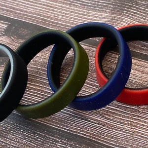 May include: Four silicone rings in different colors: black with a gray inner band, olive green, navy blue, and red. The rings are stacked on a wooden surface.