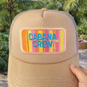 May include: Tan trucker hat with a mesh back and a curved brim. The front features a rectangular patch with the words "CABANA CREW" in blue, with a cocktail graphic. The patch has a striped background in pink, orange, and yellow.