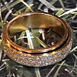 May include: A gold-toned spinning ring with a textured outer band. The ring is made of metal and has a smooth inner band.