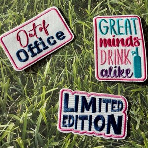 May include: Three embroidered patches with white backgrounds and pink borders are displayed on a grassy surface. The patches feature text in various colours: "Out of Office," "Great minds drink alike," and "Limited Edition."