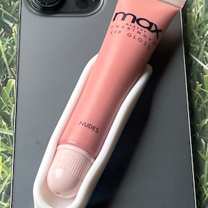Könnte beinhalten: Ein hellrosa Tube MAX Cherimoya Lipgloss, gehalten in einem weißen Halter, liegt auf einem grauen Smartphone. Auf dem Lipgloss-Tiegel steht in schwarzer Schrift "Nudes". Das Smartphone liegt auf einer grünen Oberfläche.