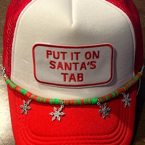 May include: Red and white trucker hat with the text "PUT IT ON SANTA'S TAB" on the front. The hat has a red mesh back and a curved brim. A beaded strand with snowflake charms is attached to the brim.