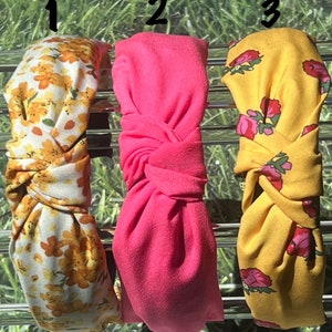 May include: Three fabric headbands. One is yellow with orange floral print, the second is solid pink, and the third is yellow with pink floral print. The headbands are knotted at the top and are displayed on a metal rack.