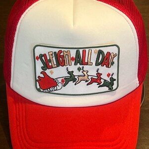 May include: A red and white trucker hat with a mesh back. The front panel features a patch with the text "SLEIGH ALL DAY" and a graphic of Santa's sleigh and reindeer. The hat has a red brim and a white front panel.