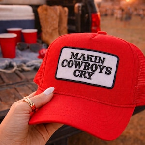 May include: A red trucker hat with a white and black patch that reads "MAKIN COWBOYS CRY." The hat has a mesh back and a curved brim. The hat is being held up by a person.