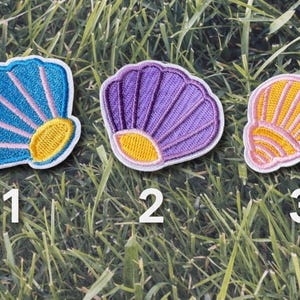 Könnte beinhalten: Drei gestickte Muschelabzeichen in Blau, Lila und Gelb mit rosa und weißen Akzenten. Jedes Abzeichen hat eine Nummer von 1 bis 3 darunter. Die Abzeichen liegen auf einer grünen Grasfläche.