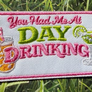 Puede incluir: Parche bordado con la frase "You Had Me At DAY DRINKING". El parche presenta ilustraciones de un cóctel y una bebida con pajita y rodaja de naranja, todo delineado en rosa, verde y blanco.