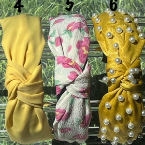 May include: Three different styles of headbands. The first is a solid yellow headband with a knot in the center. The second is a white headband with a pink floral pattern and a knot in the center. The third is a yellow headband with a velvet texture and pearl embellishments.