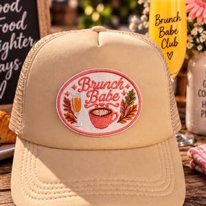 Może przedstawiać: Beżowa czapka typu trucker z owalną naszywką z wyhaftowanym różowym napisem "Brunch Babe". Naszywka przedstawia kieliszek do szampana, filiżankę kawy z wzorem latte art oraz liście w jesiennych kolorach.