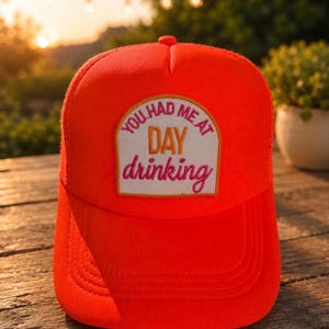 May include: Vibrant orange trucker hat with a curved peak. The hat displays a rectangular patch with the text "YOU HAD ME AT DAY drinking" in white, orange, and pink lettering. The hat is on a wooden surface.