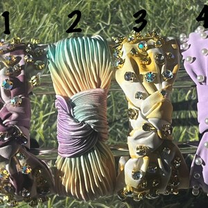 May include: Four different hair scrunchies with various embellishments. The first scrunchie is purple and white with iridescent rhinestones. The second scrunchie is a rainbow ombre with a pleated design. The third scrunchie is white and yellow with gold and iridescent rhinestones. The fourth scrunchie is lavender with white pearl embellishments.