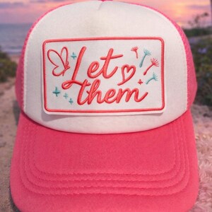 May include: A pink and white trucker hat with the phrase "Let them" embroidered in red script on a white patch. The hat features a pink brim and mesh backing, with a butterfly and floral accents.