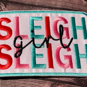 May include: Embroidered patch with the words "SLEIGH GIRL SLEIGH" in red, pink, teal, and black. The patch has a white background and a teal border. The patch is rectangular with rounded corners.