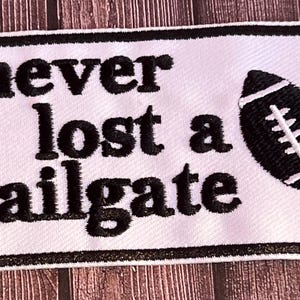 May include: White rectangular patch with black border. The text "never lost a tailgate" is embroidered in black, along with a black and white football graphic. The patch is likely for a sports fan.