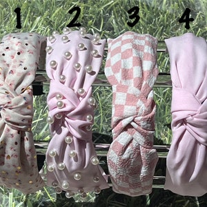May include: Four fabric headbands in a row. The first headband is white with a floral pattern and gold accents. The second is light pink with pearl embellishments. The third is pink and white checkered. The fourth is solid light pink.